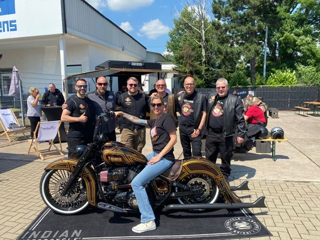 Eine Frau sitzt lächelnd auf einem kunstvoll lackierten Motorrad, umgeben von Bikern in dunkler Kleidung