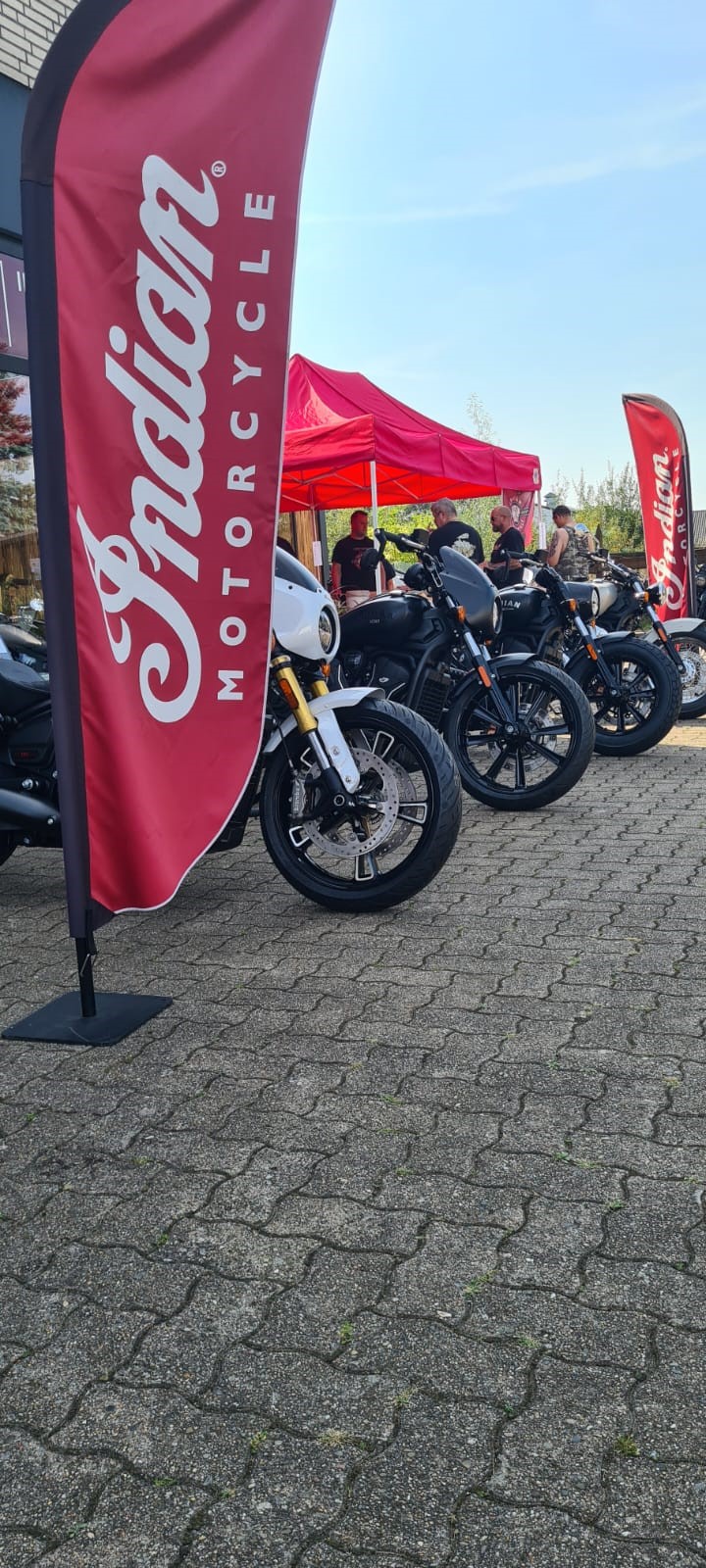 Ein Mann mit neutralem Gesichtsausdruck steht neben schwarzen Motorrädern und roten Fahnen