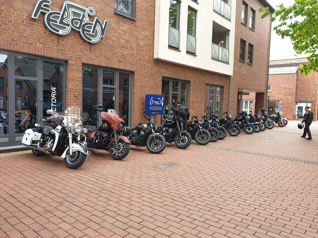 Mehrere Motorradfahrer stehen lächelnd vor roten und schwarzen Motorrädern an Gebäuden
