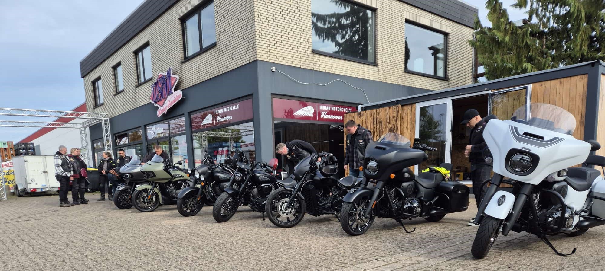 Mehrere Menschen in dunkler Kleidung stehen lachend vor schwarzen Motorrädern