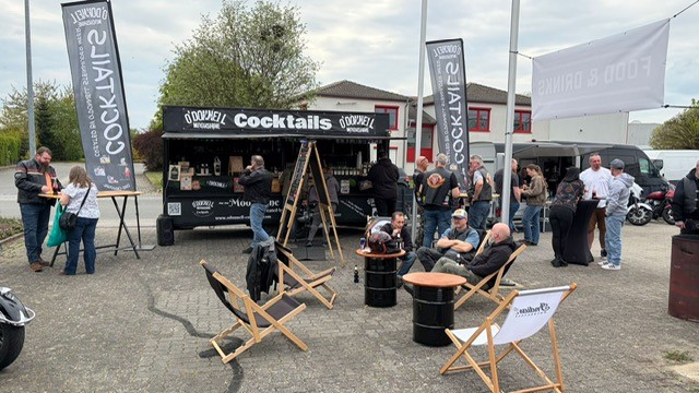 Mehrere Personen entspannen lächelnd mit Cocktails vor einem schwarzen Getränkewagen
