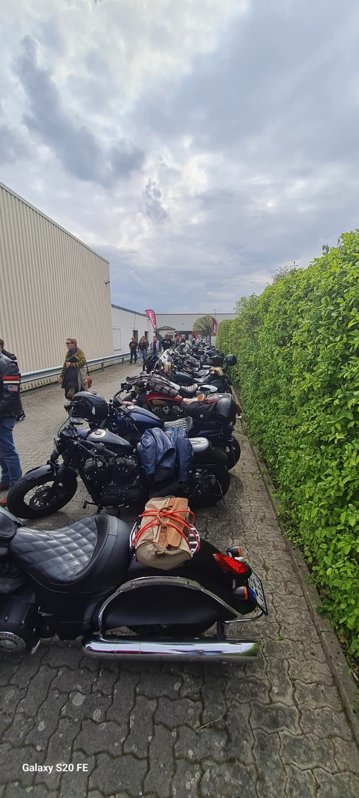 Mehrere Personen stehen freundlich an schwarzen Motorrädern neben einer grünen Hecke