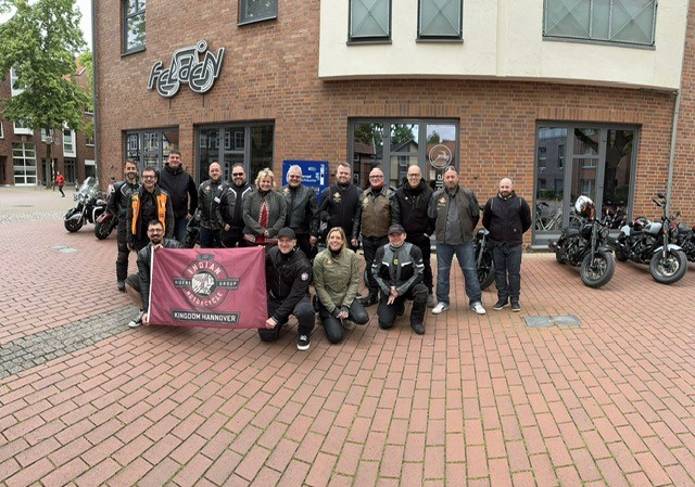 Eine Gruppe von Menschen lächelt vor einem Backsteingebäude mit Motorrädern und Fahne